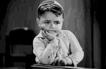 a black and white photo of a young boy sitting at a table with his hand on his chin .