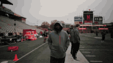 a man in a hooded jacket stands on a football field in front of a scoreboard that has the letter m on it