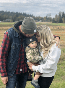 a man wearing a carhartt hat kisses a woman while holding a baby