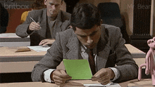 a man sits at a desk with a green piece of paper in front of him and a britbox logo in the background