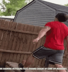 a man in a red shirt and gray shorts is jumping over a wooden fence in front of a house .