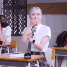 a girl in a school uniform is sitting at a desk in a classroom and covering her mouth with her hand .