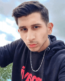 a young man wearing a black hoodie and a silver chain around his neck looks at the camera