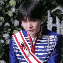 a young man wearing a blue jacket with a red and white sash around his neck is standing in front of flowers .