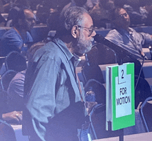 a man stands in front of a microphone behind a sign that says " for motion "