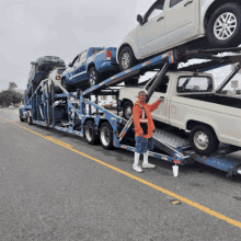 a man in an orange vest stands in front of a truck carrying cars and trucks