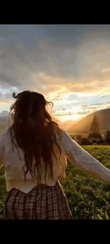 a woman is standing in a field with her arms outstretched looking at the sunset .