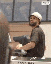 a man with a beard wearing a hat is sitting at a table in front of a sign that says back bay .