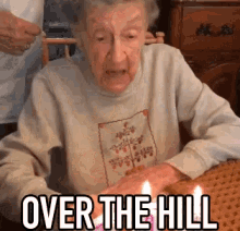 an elderly woman is sitting at a table with a cake and candles and the words over the hill above her