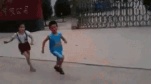 a boy and a girl are running on a sidewalk in front of a fence with chinese writing on it .