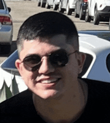 a man wearing sunglasses and a black shirt is smiling in front of a parking lot .