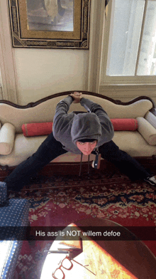 a man in a hoodie is doing a split on a couch with the caption his ass is not willem defoe