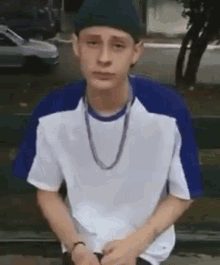 a young man wearing a hat and a necklace is sitting on a bench in front of a car .