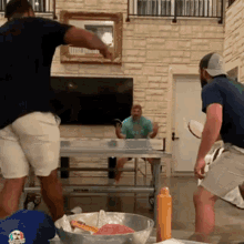 two men are playing ping pong in a living room while a man in a green shirt sits on a table