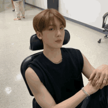 a young man in a black vest sits in a chair with his hand on his wrist