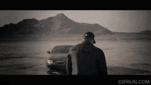 a black and white photo of a man standing in front of a bmw with a mountain in the background