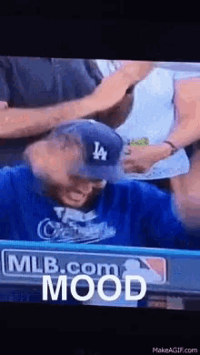 a man wearing a la hat and a blue shirt is sitting in a stadium with mlb.com written on the screen .