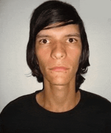 a young man with long hair is wearing a black t-shirt and looking at the camera .