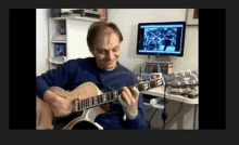 a man in a blue sweater is playing a guitar in front of a computer monitor .