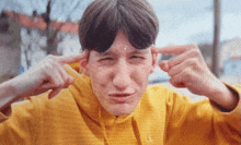 a young man in a yellow sweatshirt is covering his ears with his hands .