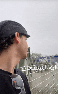 a man wearing sunglasses and a black hat stands in front of a fence with arabic writing on it