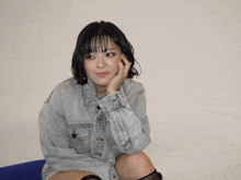 a woman in a denim jacket sits on a blue chair with her hand on her face