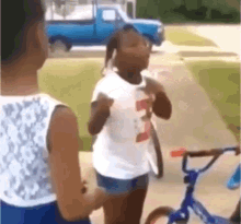 two girls are standing next to each other on a sidewalk in front of a blue truck and a bicycle .