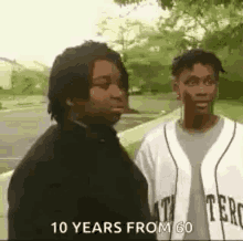 two men are standing next to each other on a sidewalk and one of them is wearing a baseball jersey .