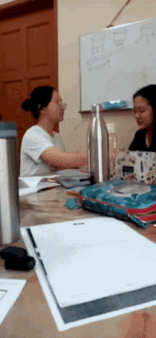 two women sit at a table in front of a white board that says ' stainless steel bottle ' on it