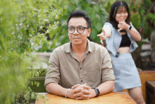 a man with glasses sits at a table with his hands folded and a woman behind him