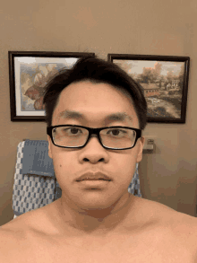 a man without a shirt is wearing glasses in front of two framed paintings