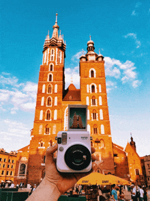 a person is holding a white instax camera in front of a large building