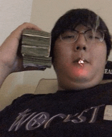 a man with glasses is smoking a cigarette while holding a stack of money in his hand .