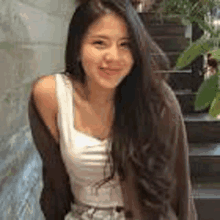 a woman with long hair is standing in front of stairs wearing a white tank top and a brown jacket .