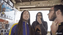 three men are standing next to each other in front of a vending machine . one of the men has dreadlocks .