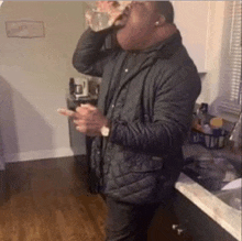 a man in a jacket is drinking water from a bottle in a kitchen .