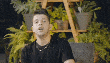 a man wearing a black shirt and a necklace is sitting in front of a shelf of potted plants .