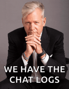 a man in a suit and tie is sitting with his hands folded in front of a sign that says we have chat logs