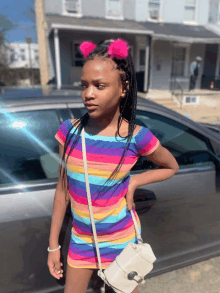 a young girl wearing a rainbow striped dress and a white purse stands in front of a car