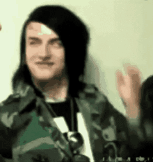 a man with long black hair is wearing a camouflage jacket and smiling while waving his hand .