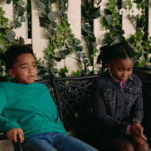 a boy in a green shirt sits next to a girl in a black jacket on a bench sponsored by nick