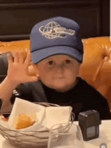 a young boy wearing a hat is sitting at a table with a basket of food .