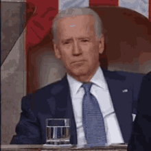 a man in a suit and tie is sitting in front of an american flag with a glass of water .