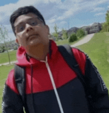 a young man wearing glasses and a red and black jacket with a backpack is standing in a park .