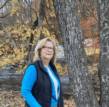 a woman wearing glasses and a blue vest stands in front of a tree