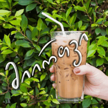 a person is holding a glass of iced coffee with a straw in front of a bush