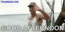 a man in a white tank top is waving at the camera while standing on a boat in the ocean .