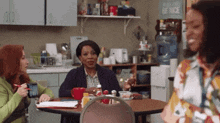 three women are sitting at a table in a kitchen with a sign on the wall that says " be nice "