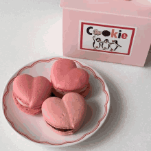 three heart shaped cookies are on a plate next to a pink cookie box