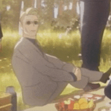 a man in a suit and sunglasses is sitting on the ground at a picnic table .